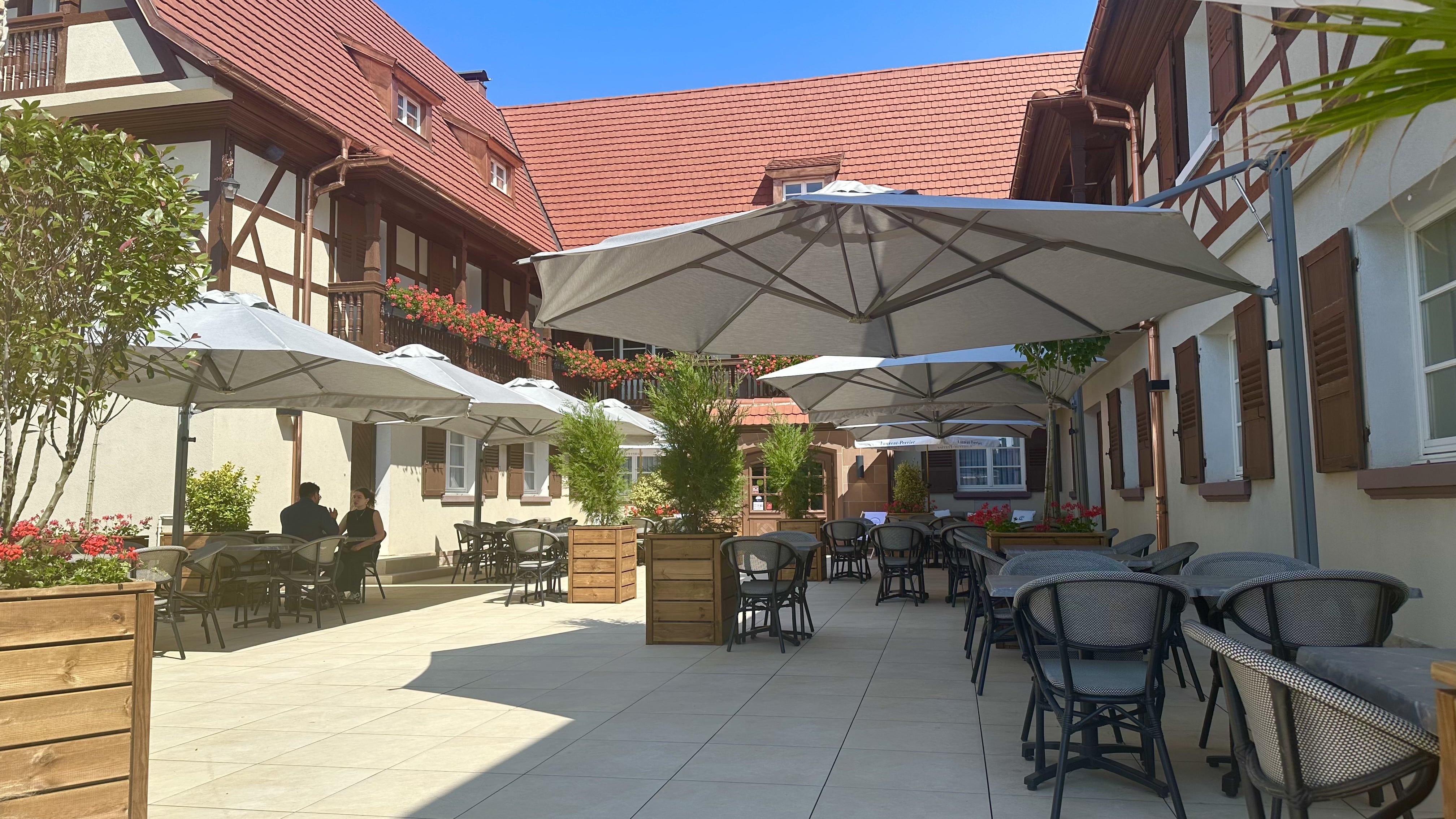 Terrasse conviviale de l’Auberge du Kochersberg, tables dressées sous parasols, façades à colombages fleuries et ambiance paisible en Alsace.