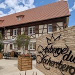 Entrée de l’Auberge du Kochersberg : mur de pierre avec logo et cour fleurie, maison à colombages typique sous le ciel bleu.